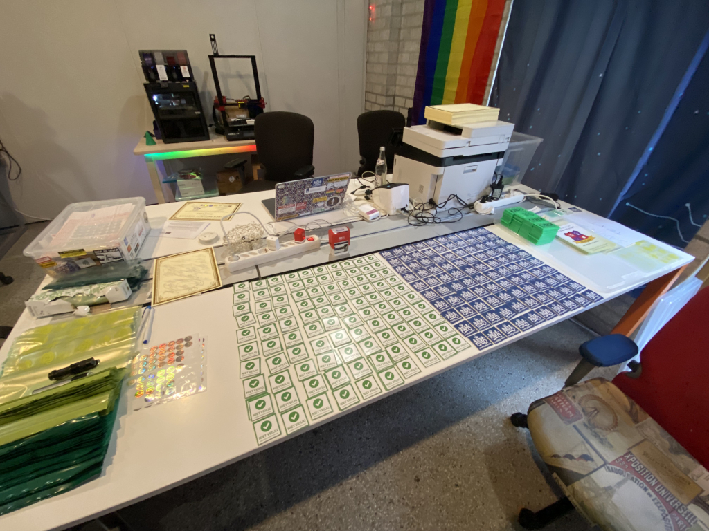 De certificaatprinter met een stapel certificaten en stapels stickers om te verzenden. Dit toont een tafel met daarop een productiestraat om enveloppen te vullen met allerlei items. Deze foto is genomen bij hackerspace hackalot in eindhoven.